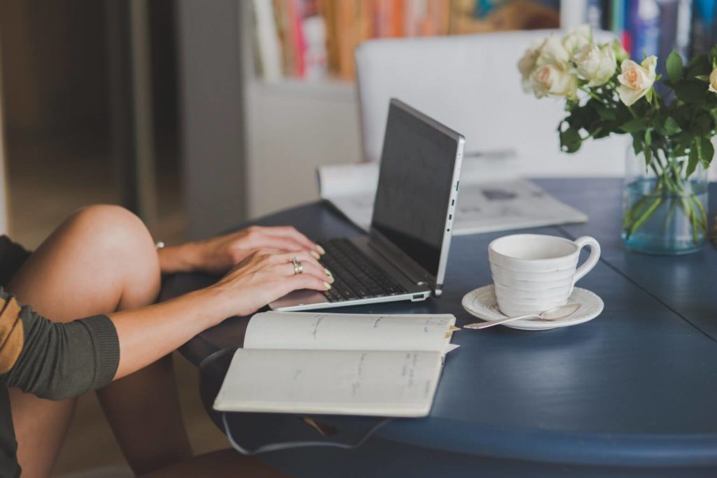 Image montrant de côté un ordinateur portable avec une tasse de café et un carnet ouvert à côté/ Une femme a les mains posées sur le clavier