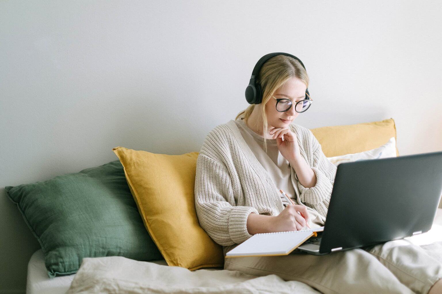 Une jeune femme blonde assise sur un canapé avec un casque audio sur les oreilles et qui travaille sur son ordinateur portable